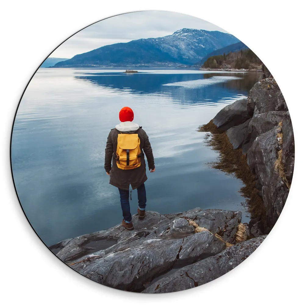 Dibond Muurcirkel - Man met Gele Rugtas aan de Rand van Meer in Landschap vol Bergen